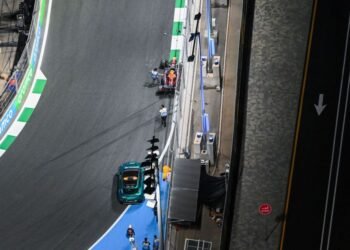 Vista aérea de uma pista de corrida com um carro parado, equipes próximas ao veículo e sinalização de trânsito visível.