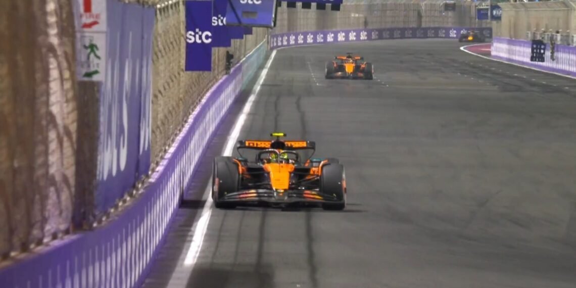 Carro de corrida laranja em alta velocidade em pista urbana, seguido por outro carro. Corrida noturna de Fórmula 1.