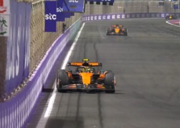 Carro de corrida laranja em alta velocidade em pista urbana, seguido por outro carro. Corrida noturna de Fórmula 1.