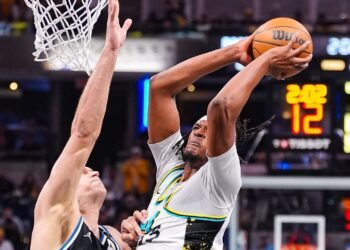 Jogador de basquete executa uma enterrada sob defesa durante uma partida intensa, capturando o dinamismo do esporte.