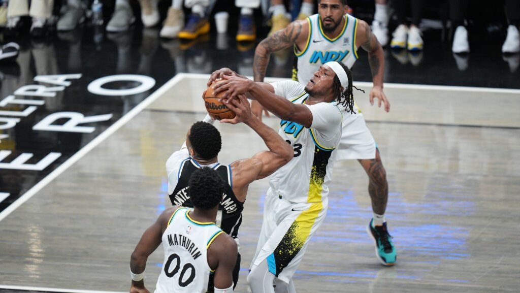 Jogadores de basquete competem por posse de bola durante partida intensa na quadra, com público atento ao fundo.