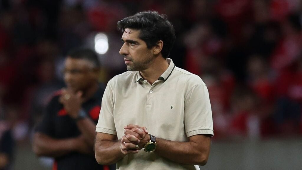 Treinador de futebol, concentrado, veste camisa polo bege no estádio lotado. Ambiente de competição intensa.