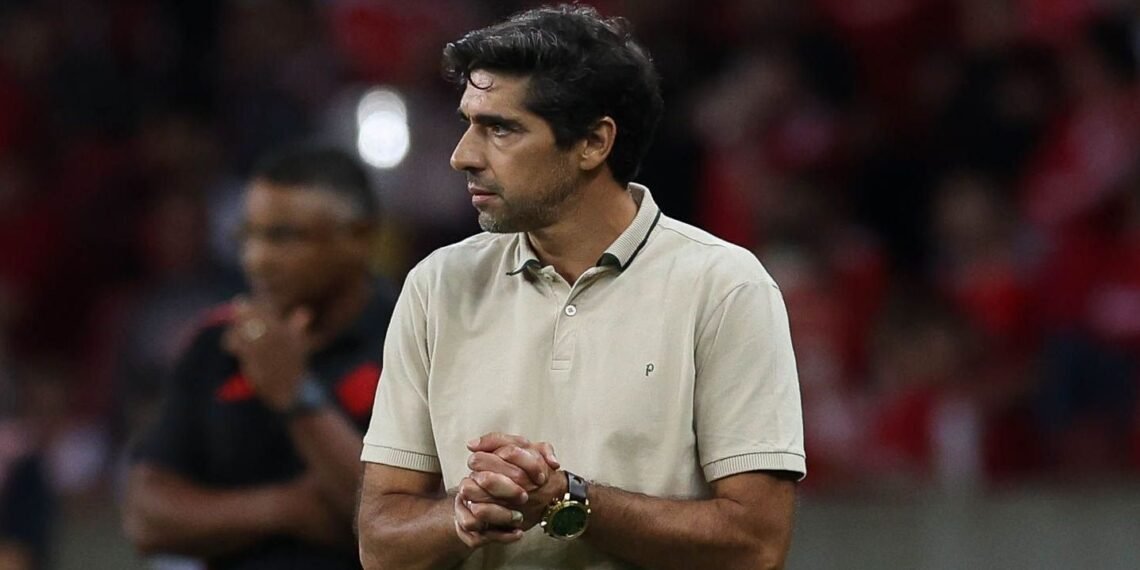 Treinador de futebol, concentrado, veste camisa polo bege no estádio lotado. Ambiente de competição intensa.