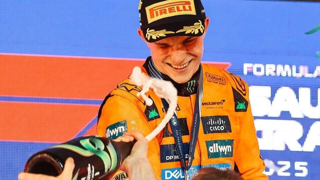 Piloto de Fórmula 1 celebra no pódio com champanhe, sorrindo, vestindo uniforme laranja da equipe McLaren.
