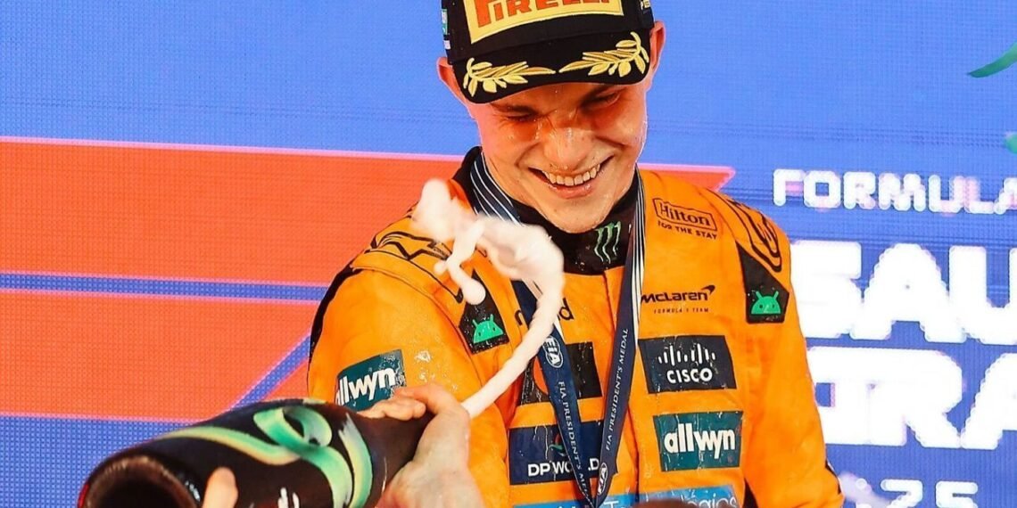Piloto de Fórmula 1 celebra no pódio com champanhe, sorrindo, vestindo uniforme laranja da equipe McLaren.
