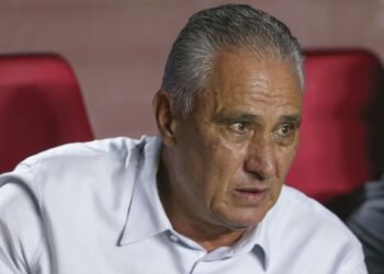 Presidente revela negociações com Tite no Corinthians: 'Era certo' 34 Homem de camisa branca observa atentamente algo fora do quadro, sentado em uma cadeira vermelha, em ambiente interno.