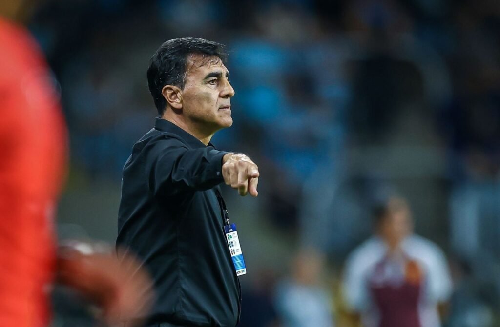 Treinador de futebol gesticula instruções à beira do campo durante partida. Atuações esportivas e táticas em destaque.