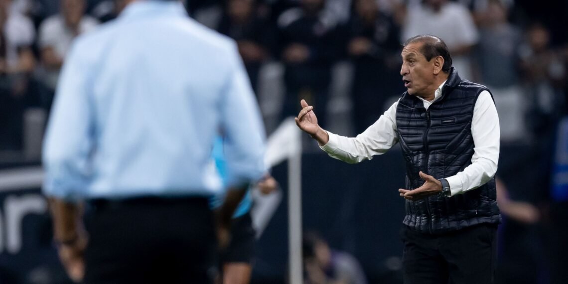Técnico de futebol gesticula energicamente à beira do campo, interagindo com outro integrante de equipe durante uma partida.