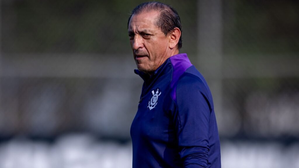 Treinador de futebol em campo, usando agasalho com emblema de time, focado e observando o ambiente ao ar livre.
