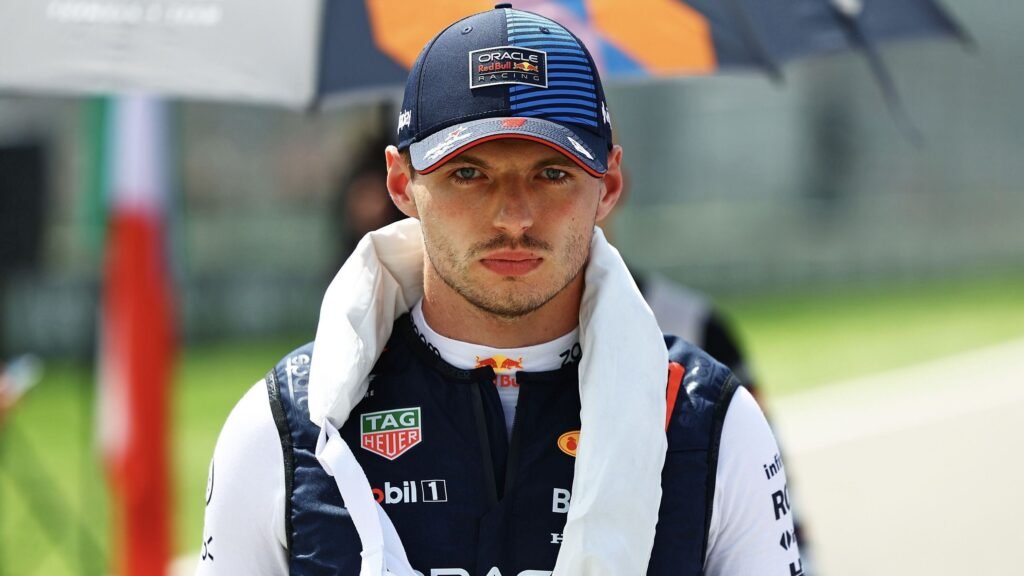 Piloto de corrida com boné azul da Oracle Red Bull Racing, veste macacão de corrida, em cenário de pista automobilística.