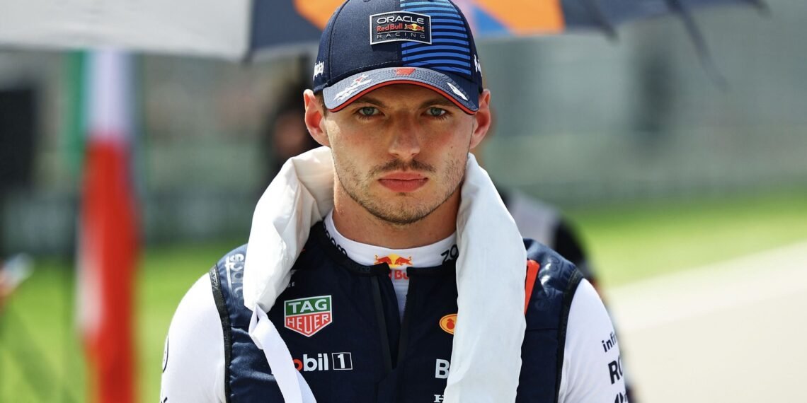 Piloto de corrida com boné azul da Oracle Red Bull Racing, veste macacão de corrida, em cenário de pista automobilística.