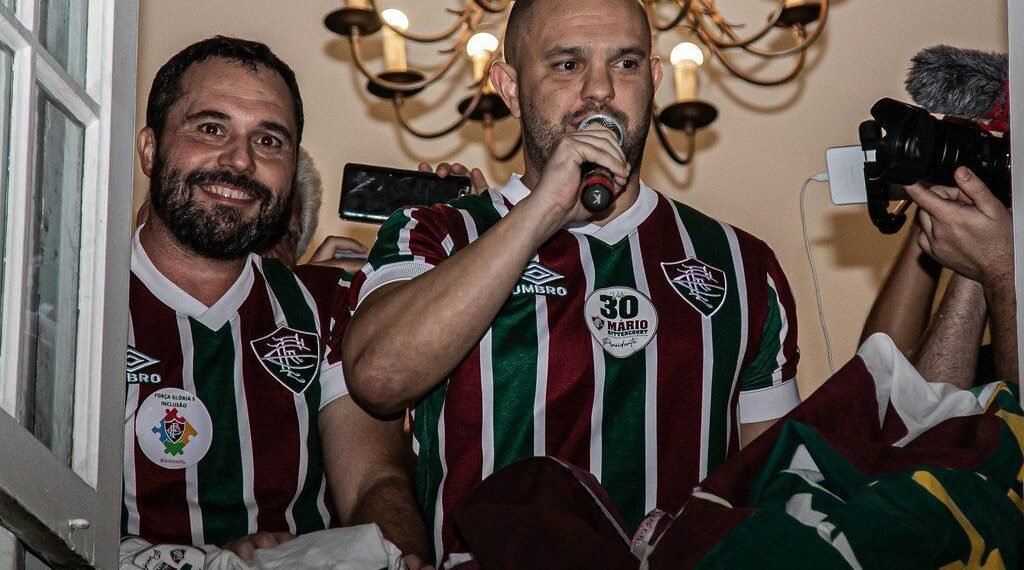 Homens com camisas do Fluminense discursam em ambiente coberto, segurando microfone, cercados por câmeras e pessoas.