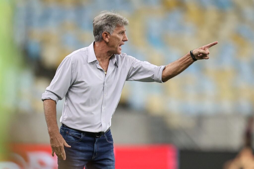 Técnico de futebol gesticula intensamente à beira do campo, vestindo camisa social e jeans, em estádio com arquibancadas desfocadas.