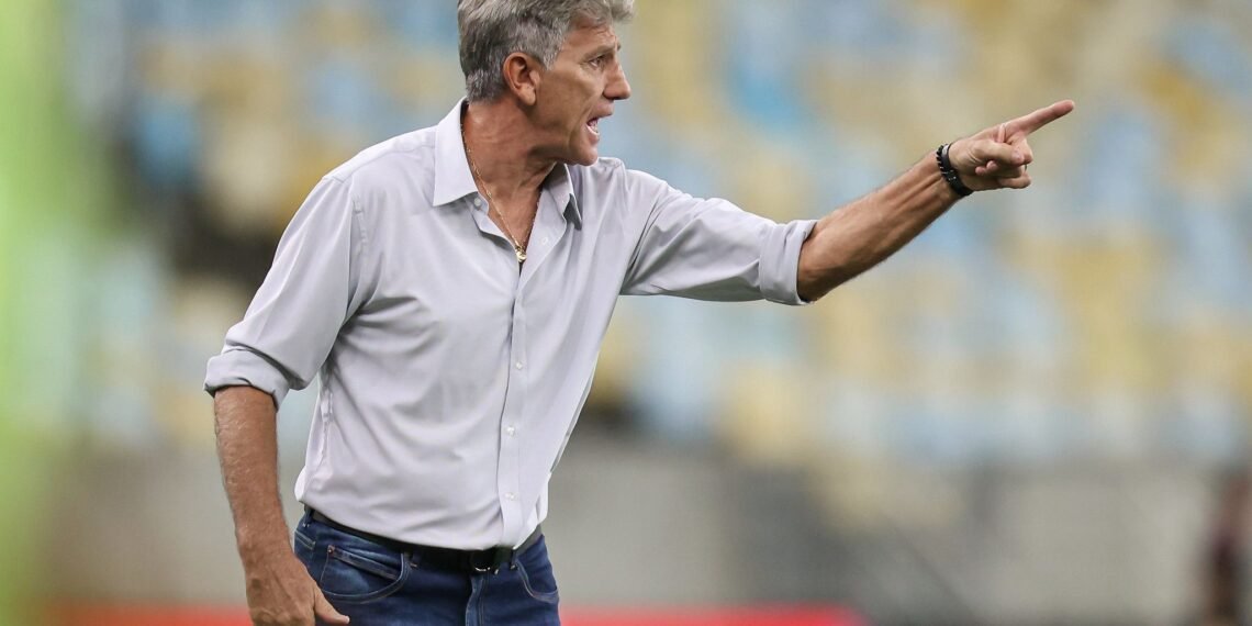Técnico de futebol gesticula intensamente à beira do campo, vestindo camisa social e jeans, em estádio com arquibancadas desfocadas.