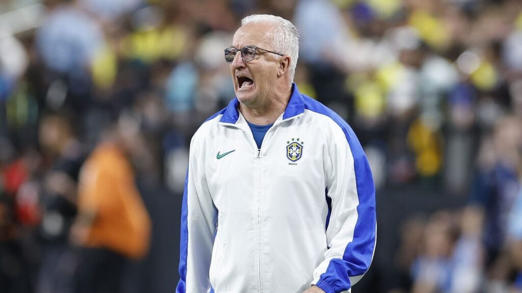 Treinador da seleção brasileira de futebol com expressão intensa, vestindo jaqueta branca no campo de jogo.
