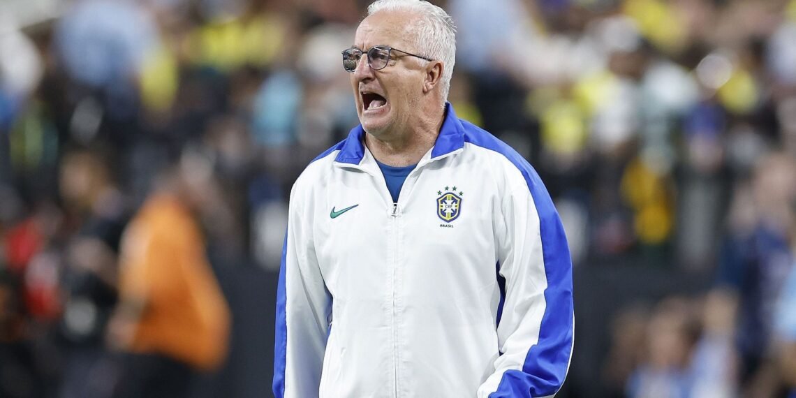 Treinador da seleção brasileira de futebol com expressão intensa, vestindo jaqueta branca no campo de jogo.