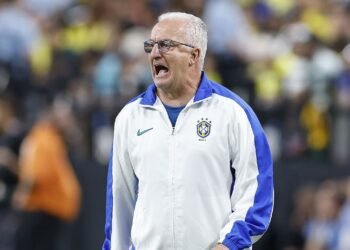 Treinador da seleção brasileira de futebol com expressão intensa, vestindo jaqueta branca no campo de jogo.