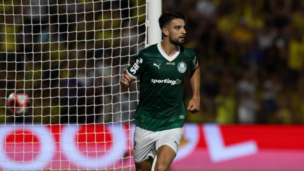 Jogador de futebol com uniforme verde correndo em campo, próximo ao gol, durante partida intensa. Torcida ao fundo.