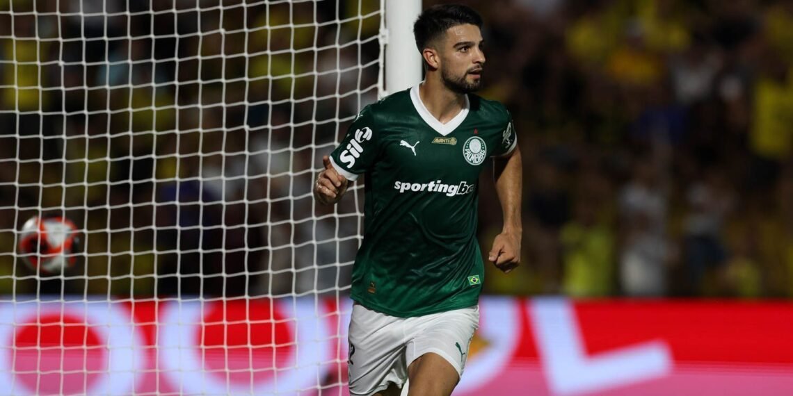 Jogador de futebol com uniforme verde correndo em campo, próximo ao gol, durante partida intensa. Torcida ao fundo.