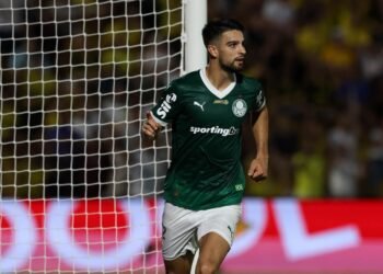Jogador de futebol com uniforme verde correndo em campo, próximo ao gol, durante partida intensa. Torcida ao fundo.