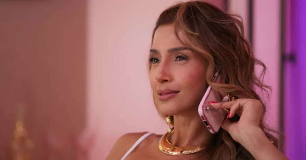 Mulher sorridente em conversa ao celular rosa, com cabelo ondulado, em ambiente iluminado. Vestida elegantemente, transmite confiança.