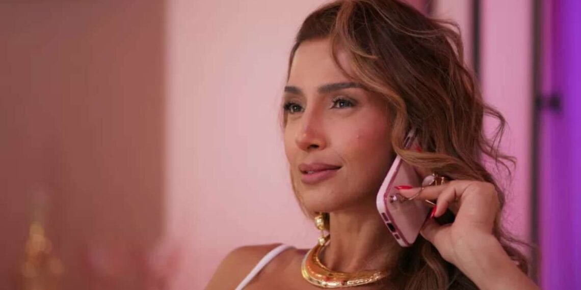 Mulher sorridente em conversa ao celular rosa, com cabelo ondulado, em ambiente iluminado. Vestida elegantemente, transmite confiança.