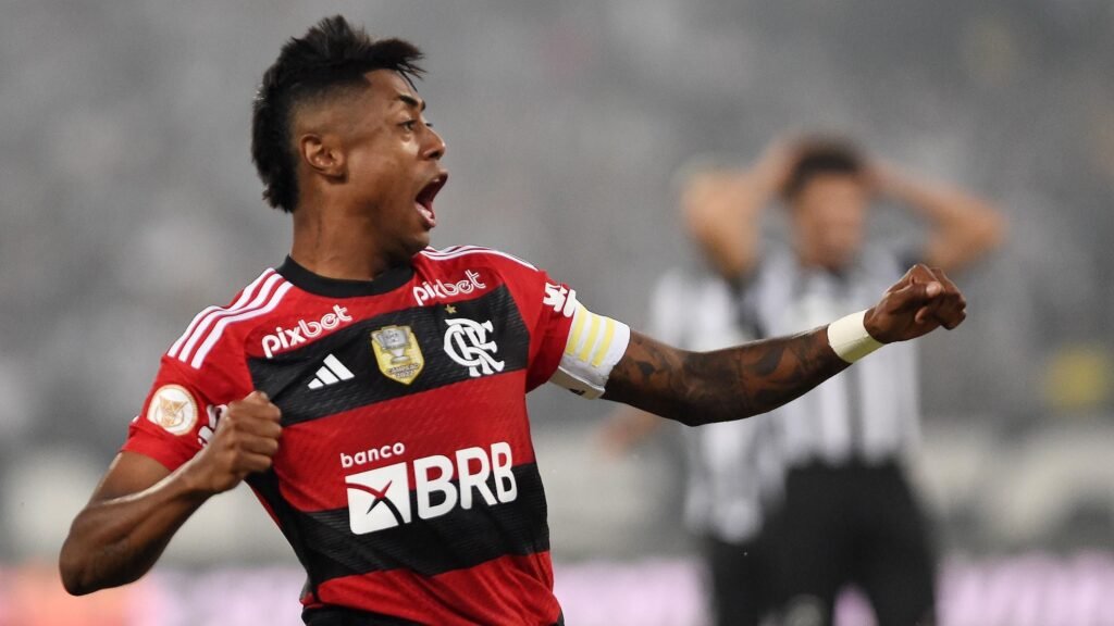 Jogador de futebol com uniforme do Flamengo comemora com o braço erguido em campo, bandeira no fundo desfocada.
