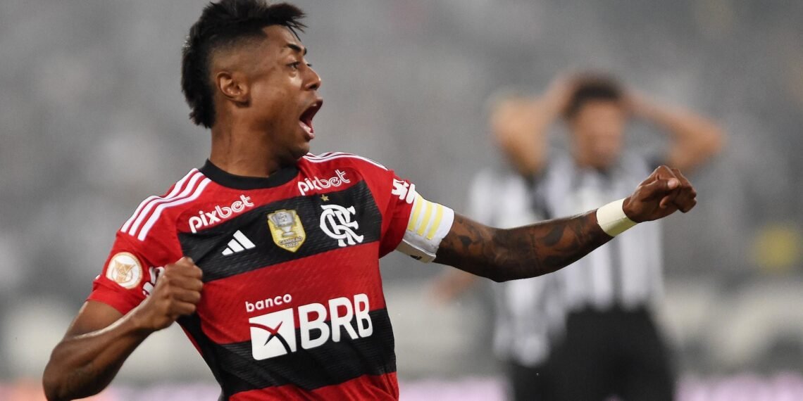 Jogador de futebol com uniforme do Flamengo comemora com o braço erguido em campo, bandeira no fundo desfocada.
