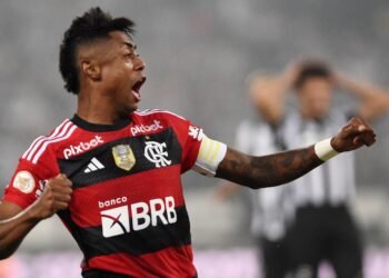 STJD solicita PF para compartilhar provas de Bruno Henrique 36 Jogador de futebol com uniforme do Flamengo comemora com o braço erguido em campo, bandeira no fundo desfocada.