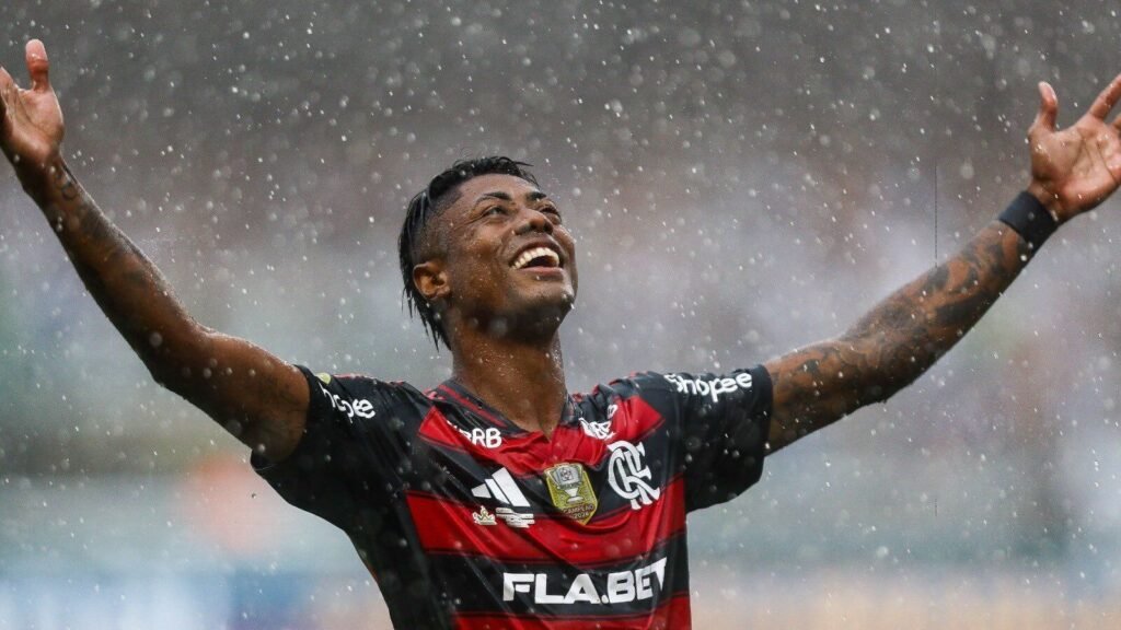 Jogador do Flamengo com expressão de vitória em campo sob chuva, braços erguidos, transmitindo emoção e energia do futebol.