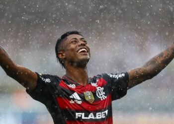 Salário de Bruno Henrique, indiciado pela PF, é revelado! 37 Jogador do Flamengo com expressão de vitória em campo sob chuva, braços erguidos, transmitindo emoção e energia do futebol.