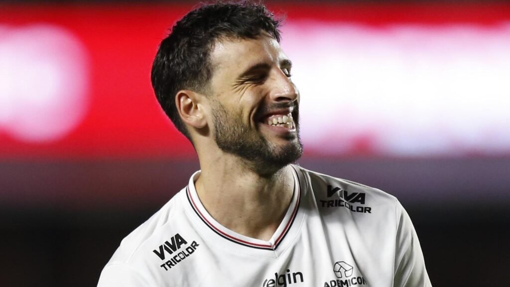 Homem sorrindo em uniforme branco de futebol, em campo iluminado, transmitindo felicidade durante uma partida noturna.