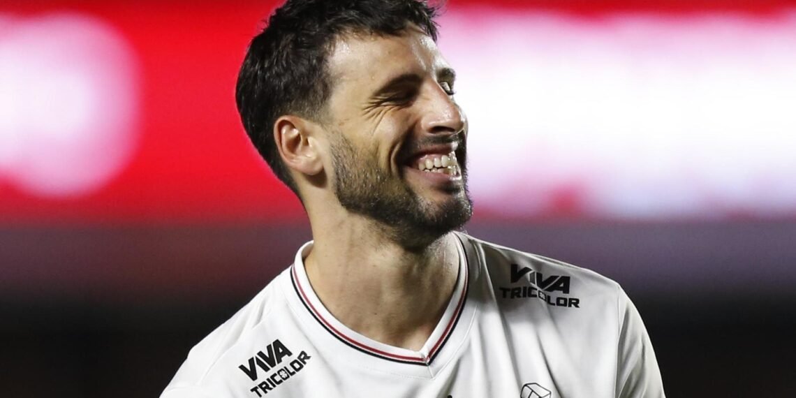 Homem sorrindo em uniforme branco de futebol, em campo iluminado, transmitindo felicidade durante uma partida noturna.