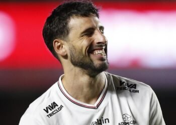 Homem sorrindo em uniforme branco de futebol, em campo iluminado, transmitindo felicidade durante uma partida noturna.