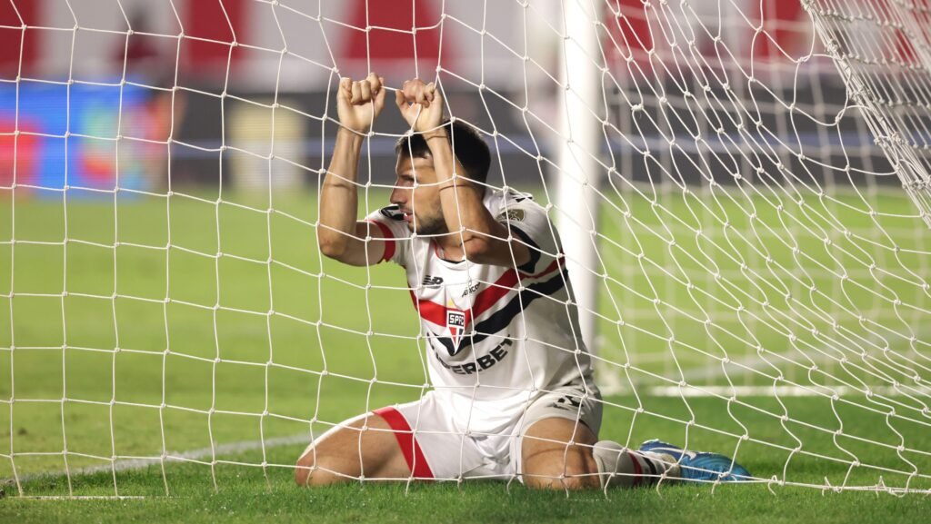 Jogador de futebol ajoelhado na área do gol, segurando a rede com expressão de frustração após jogada perdida.