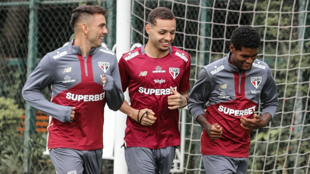 Jogadores do São Paulo FC sorridentes durante treino, vestindo uniforme do time, em campo com rede de fundo.