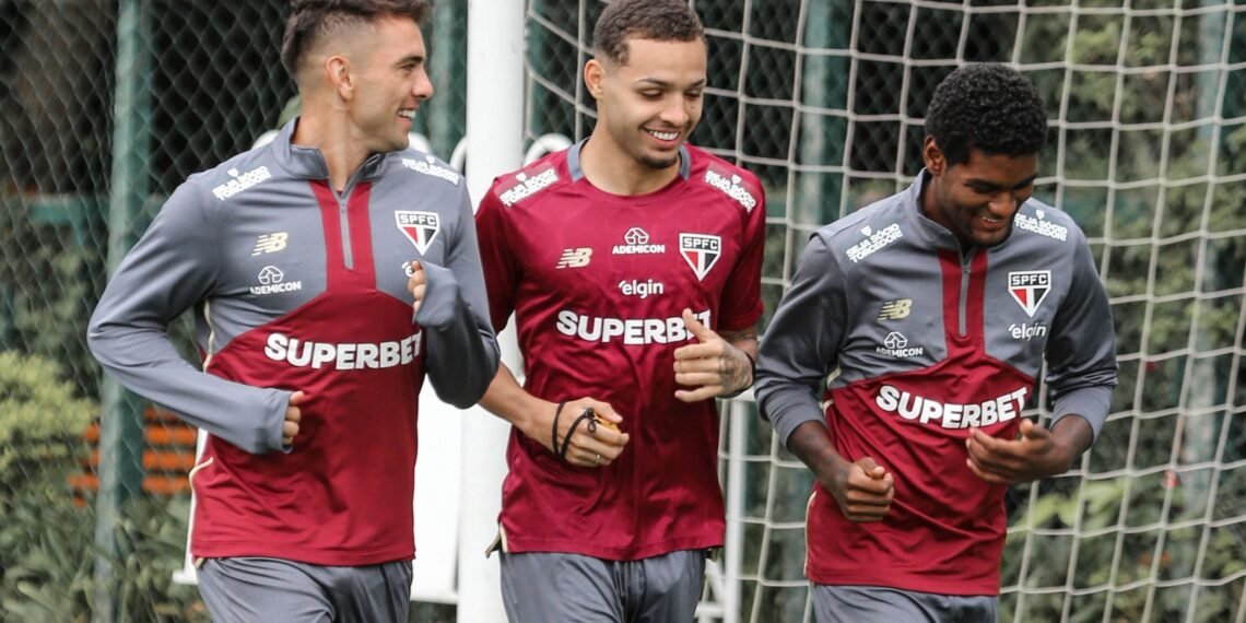 Jogadores do São Paulo FC sorridentes durante treino, vestindo uniforme do time, em campo com rede de fundo.