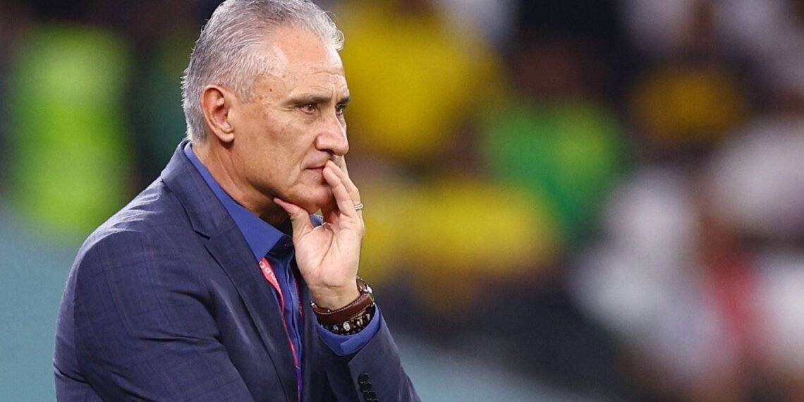 Homem pensativo com terno azul, mão no queixo, em ambiente de estádio esportivo com fundo desfocado de torcedores.
