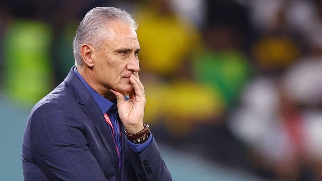 Um treinador de futebol em traje formal observa atentamente um jogo, com expressão pensativa e o estádio ao fundo.