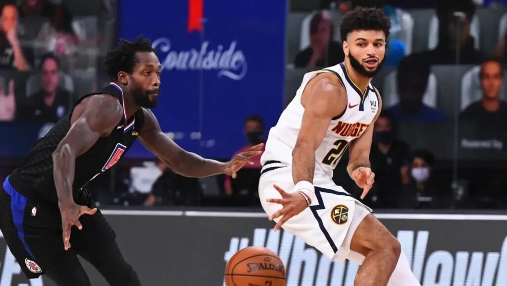 Jogador de basquete em ação desviando de marcador em partida, vestindo uniformes das equipes, com foco na bola.