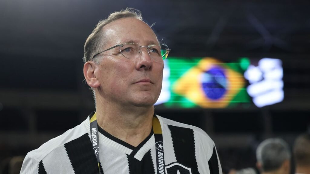 Homem de óculos com camisa listrada preto e branco assiste evento esportivo; bandeira do Brasil ao fundo.