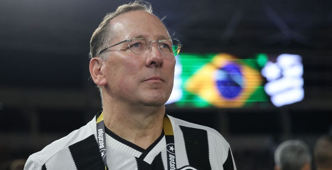 Homem de óculos com camisa listrada preto e branco assiste evento esportivo; bandeira do Brasil ao fundo.