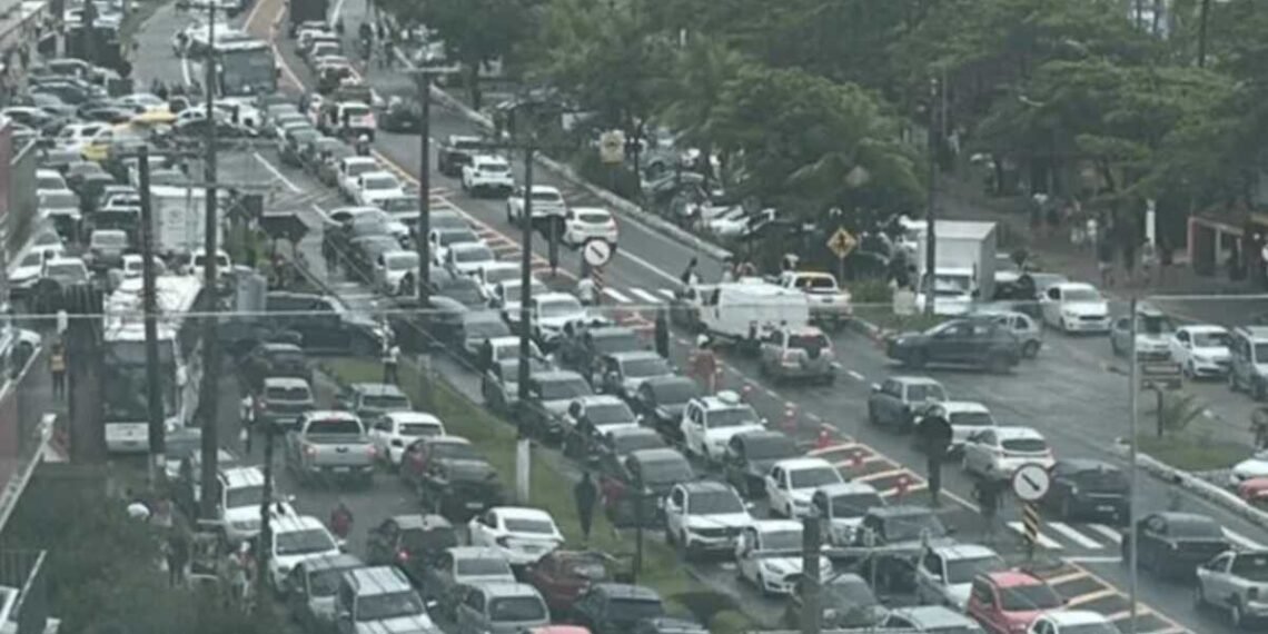 Trânsito congestionado em avenida movimentada, com longas filas de carros parados, árvores e pedestres ao fundo.