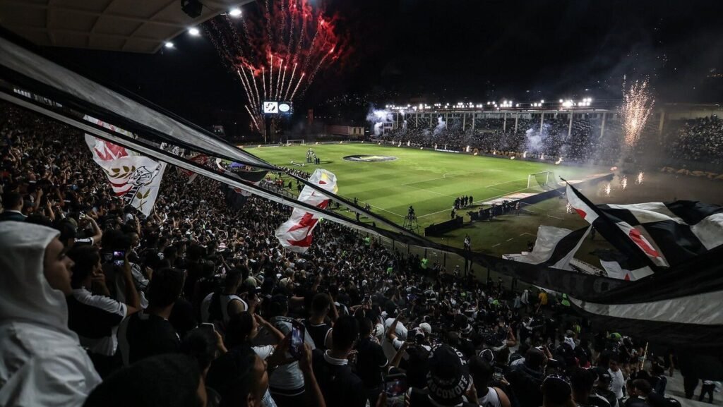 Estádio lotado com torcedores animados empunhando bandeiras, fogos de artifício iluminam o céu durante jogo de futebol.