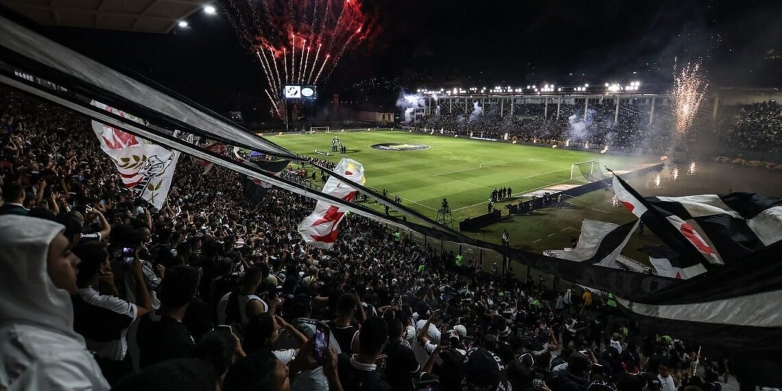 Estádio lotado com torcedores animados empunhando bandeiras, fogos de artifício iluminam o céu durante jogo de futebol.