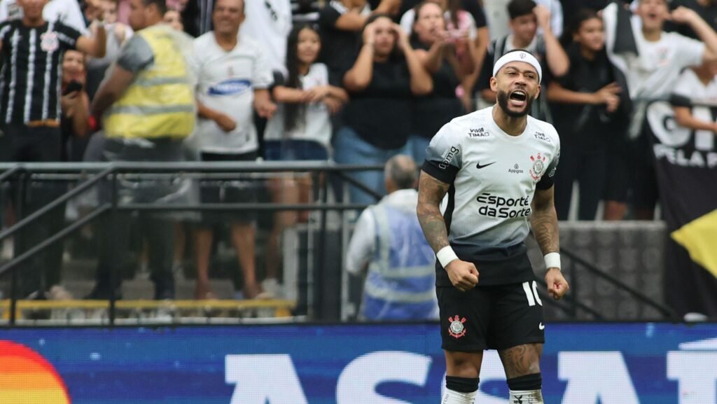 Jogador do Corinthians comemorando gol com expressão de vitória; torcida ao fundo vibra intensamente no estádio.