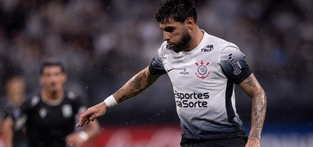 Jogador de futebol do Corinthians conduz a bola durante partida, com foco e determinação no campo de jogo molhado.