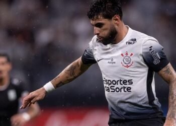 Jogador de futebol do Corinthians conduz a bola durante partida, com foco e determinação no campo de jogo molhado.