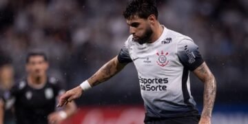 Jogador de futebol do Corinthians conduz a bola durante partida, com foco e determinação no campo de jogo molhado.