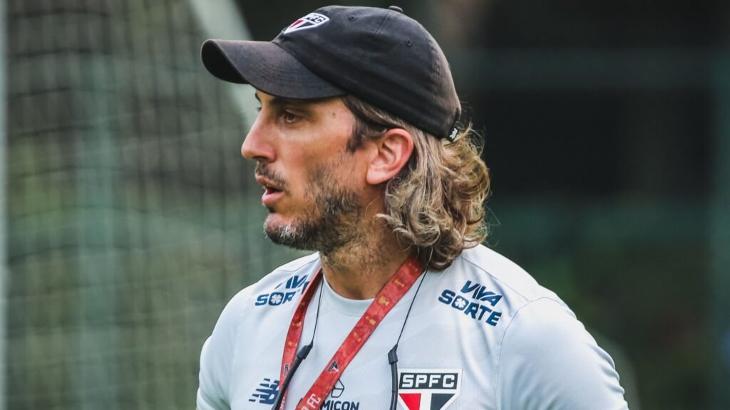 Homem com boné preto e camisa do São Paulo FC no campo, aparentando concentração. Fundo desfocado de rede de futebol.